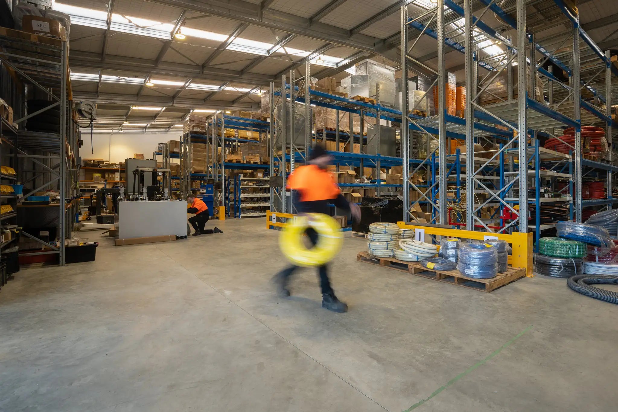 An image of the United Flexible Interior of a their busy warehouse with employees at work, featuring high storage racks filled with materials and a worker in motion carrying a yellow coil of hose, highlighting the dynamic industrial environment.