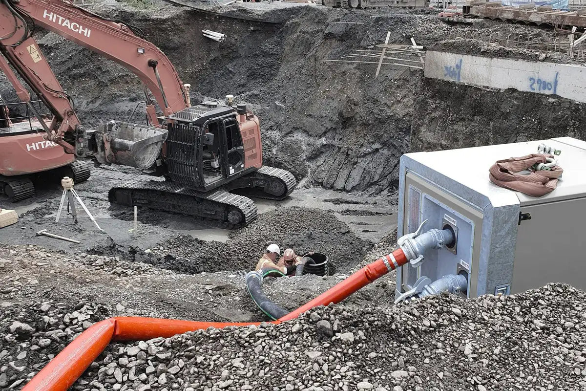 Construction site with workers installing underground utilities near an excavator and a large concrete vault. Features include heavy machinery, large hoses, and rocky terrain, reflecting an active excavation and installation scene.