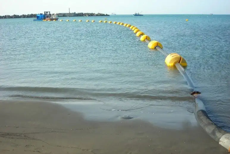 Large industrial hose with yellow buoys extending into the sea from a sandy beach, used for maritime operations, set against a tranquil blue water backdrop with a boat visible in the distance.