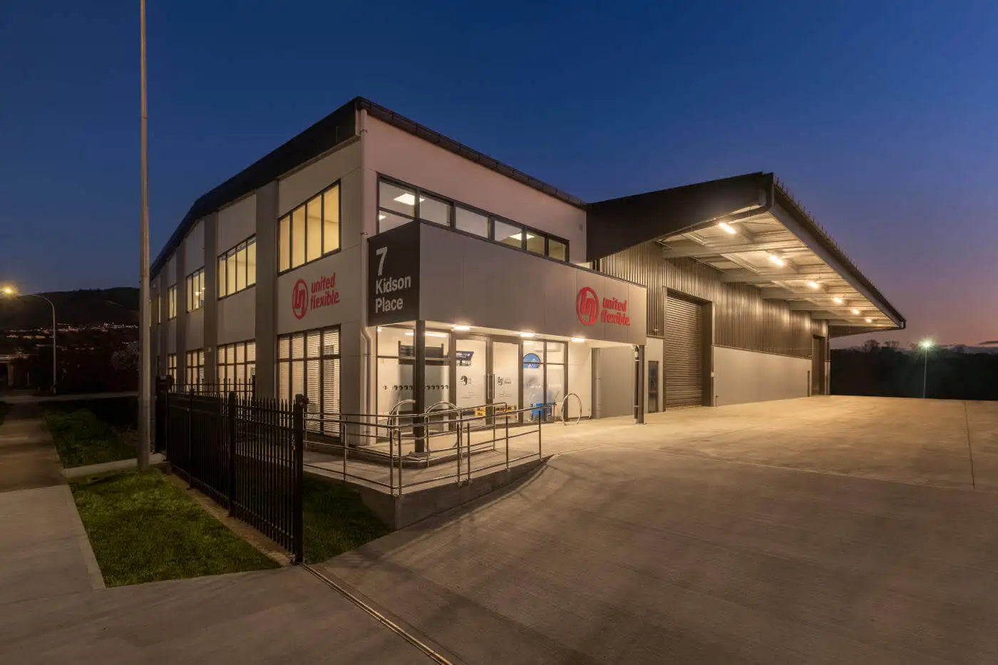 Exterior of a modern industrial building at twilight, featuring a two-story structure with illuminated signage and a large warehouse section, all under a clear evening sky.