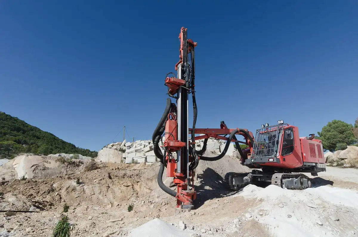 Large red drilling rig on a construction site with clear blue skies, operating on rocky terrain in a mountainous area, emphasizing heavy machinery in industrial and geological work settings.